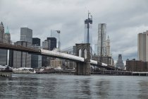 Desde debajo del puente de Manhattan visto desde la orilla del río con edificios de la ciudad y cielo gris nublado en el fondo en la ciudad de Nueva York - foto de stock