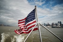 National flag of USA waving on pole of floating vessel against cloudy sky near New York City coast — Stock Photo