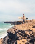 Vue latérale d'un homme torse nu avec planche de surf debout sur une côte pierreuse et admirant la mer ondulante sur l'île de Fuerteventura, Espagne — Photo de stock