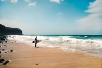 Homem anônimo de corpo inteiro com prancha de surf em pé na praia de areia perto da água do mar acenando no dia ensolarado na Ilha Fuerteventura, Espanha — Fotografia de Stock