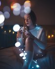 Stylish young female in sweater and socks sitting on bed and examining glowing fairy lights in cozy dark room at home — Stock Photo
