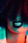 Crop female model in synthetic wig and with black lipstick showing teeth under colorful light — Stock Photo