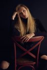 Jeune femme blonde en vêtements sombres assis sur une chaise sur fond noir et regardant ailleurs — Photo de stock