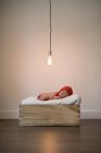 Чарівна дитина в червоному капелюсі лежить на м'якій ковдрі і спить в дерев'яній коробці під блискучою лампою — стокове фото
