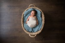 Вид сверху новорожденного, завернутого в ткань, лежащего на мягком одеяле и спящего в плетеной корзине на полу дома — стоковое фото