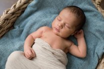 Вид сверху новорожденного, завернутого в ткань, лежащего на мягком одеяле и спящего в плетеной корзине на полу дома — стоковое фото