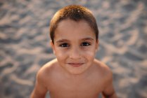 Joyeux garçon torse nu souriant et regardant la caméra tout en se tenant sur la plage de sable pendant le coucher du soleil — Photo de stock
