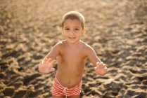 Счастливый мальчик без рубашки улыбается и смотрит в камеру, стоя на песчаном пляже во время заката — стоковое фото