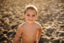 Feliz chico sin camisa sonriendo y mirando a la cámara mientras está de pie en la playa de arena durante la puesta del sol - foto de stock