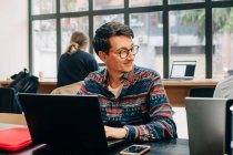 Молодой сконцентрированный мужчина в повседневной одежде и очках сидит за столом и печатает на ноутбуке во время работы над проектом в креативном рабочем пространстве — стоковое фото