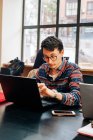 Молодий концентрований чоловік у повсякденному вбранні та окулярах сидить за столом і друкує на ноутбуці під час роботи над проектом у творчій робочій області — стокове фото