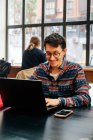 Jovem macho concentrado em roupa casual e óculos sentados na mesa e digitando no laptop enquanto trabalhava no projeto no espaço de trabalho criativo — Fotografia de Stock