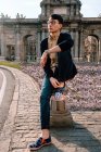 Ganzkörper asiatischer junger trendiger Mann in stylischem Outfit und Brille, der auf einer kleinen Steinsäule sitzt und in der Nähe eines schönen alten Gebäudes auf der Straße wegschaut — Stockfoto