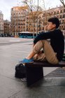 Ganzkörper asiatischer junger trendiger Mann in stylischem Outfit und Brille, der auf einer kleinen Steinsäule sitzt und in der Nähe eines schönen alten Gebäudes auf der Straße wegschaut — Stockfoto