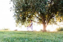 Vista lateral do macho distância em roupas brancas de pé em um joelho enquanto faz ioga e meditando no gramado verde contra a árvore na noite de verão no parque — Fotografia de Stock