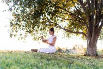 Seitenansicht einer ruhigen jungen Frau in weißer Kleidung, die auf Knien sitzt und die tibetische Klangschale in den Händen hält, während sie an Sommertagen Yoga macht und sich auf dem Rasen entspannt — Stockfoto