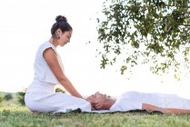 Calm couple meditating while man lying on mat with eyes closed and woman sitting on knees and holding partner by head during looking down in green meadow — Stock Photo