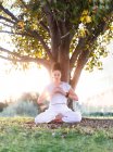 Side view of calm young female in white wear sitting on knees and holding Tibetan singing bowl in hands while going yoga and relaxing on lawn in summer day — Stock Photo