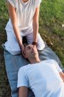 From above of calm couple meditating while man lying on mat with eyes closed and woman sitting on knees and holding partner by head during looking down in green meadow — Stock Photo