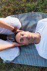 Casal calmo meditando enquanto o homem deitado no tapete com os olhos fechados e mulher sentada de joelhos e segurando parceiro pela cabeça durante o olhar para baixo no prado verde — Fotografia de Stock