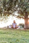 Вид збоку чоловік у білому одязі в положенні лотоса медитує на зеленому газоні проти дерева в літній вечір у парку — стокове фото
