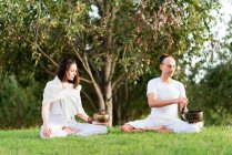 Flexible relaxed couple in white wear sitting in lotus pose holding tibetan singing bowl in hand while meditating and enjoying time together on summer day in park — Stock Photo