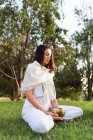 Vista laterale di calma giovane femmina in bianco indossare seduto sulle ginocchia e tenendo tibetano canto ciotola in mano mentre si va yoga e relax sul prato in giorno d'estate — Foto stock