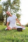 Vue latérale du jeune homme calme en tenue blanche assis sur les genoux et tenant bol de chant tibétain dans les mains tout en allant yoga et détente sur la pelouse dans la journée d'été — Photo de stock