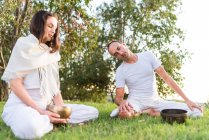 Flexible relaxed couple in white wear sitting in lotus pose with eyes closed and holding tibetan singing bowl in hand while meditating and enjoying time together on summer day in park — Stock Photo