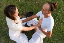 From above of calm couple in white clothes sitting opposite each other in lotus position on grass and keeping hands on stomachs and on each others hearts while meditating outdoors with singing bowls in park — Stock Photo