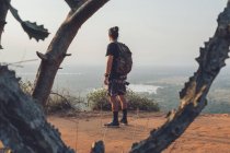 Back view of young unrecognizable male traveler in casual clothes with haversack standing on edge of cliff against majestic landscape of green forest and cloudless blue sky during sunset — Stock Photo