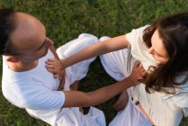From above of calm couple in white clothes sitting opposite each other in lotus position on grass and keeping hands on stomachs and on each others hearts while meditating outdoors with singing bowls in park — Stock Photo