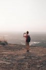 Обратный вид на молодого неузнаваемого мужчину-путешественника в повседневной одежде, стоящего на краю скалы и фотографирующего величественный ландшафт зеленого леса на фоне безоблачного голубого неба на закате — стоковое фото