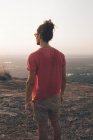 Back view of young man in casual outfit standing on rocky cliff and admiring incredible view against cloudless sky during sunset — Stock Photo