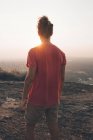 Back view of young man in casual outfit standing on rocky cliff and admiring incredible view against cloudless sky during sunset — Stock Photo