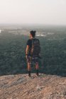 Обратный вид на молодого неузнаваемого мужчину-путешественника в повседневной одежде, стоящего на краю скалы против величественного ландшафта зеленого леса и безоблачного голубого неба на закате — стоковое фото
