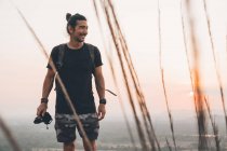 Молодий веселий чоловік мандрівник у повсякденному одязі з фотоапаратом, що стоїть на краю скелі і фотографує величний пейзаж зеленого лісу під час заходу сонця — стокове фото