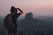 Обратный вид на молодого неузнаваемого мужчину-путешественника в повседневной одежде, стоящего на краю скалы и фотографирующего величественный ландшафт зеленого леса на фоне безоблачного голубого неба на закате — стоковое фото