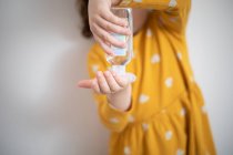 Crop little girl wearing yellow colorful dress standing in medical room and pouring antiseptic liquid from plastic bottle on hands — Stock Photo