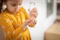 Serious little girl wearing yellow colorful dress standing in medical room and pouring antiseptic liquid from plastic bottle on hands — Stock Photo