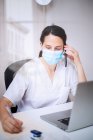 Serious young female doctor wearing white uniform and medical mask working on laptop in latex gloves sitting at desk in modern clinic — Stock Photo