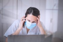 Médico femenino joven serio que usa uniforme blanco y máscara médica que trabaja en el ordenador portátil en guantes de látex sentado en el escritorio en la clínica moderna - foto de stock