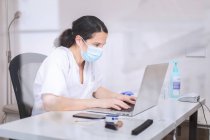 Серйозна молода жінка-лікар в білій формі та медична маска, що працює на ноутбуці в латексі, сидить за столом у сучасній клініці — стокове фото