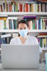 Серьёзная молодая женщина-врач в белой униформе и медицинской маске работает на ноутбуке в латексных перчатках, сидя за столом в современной клинике — стоковое фото