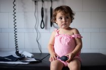Цікава чарівна маленька дівчинка в повсякденній рожевій сукні сидить на лавці в медичній кімнаті і грає з тонометром під час відвідування лікаря — стокове фото
