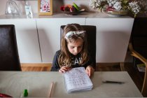 Сверху сосредоточенная девушка сидит за столом и читает заметки в блокноте, выполняя домашнее задание — стоковое фото