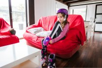 Girl in colorful helmet sitting on couch at home and putting on knee cap before riding roller skates — Stock Photo