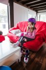 Girl in colorful helmet sitting on couch at home and putting on knee cap before riding roller skates — Stock Photo