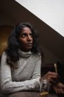 Side view of calm young Indian woman in casual dress resting on comfortable sofa near window in modern attic room while rolling up marijuana cigaret — Stock Photo