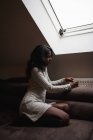 Vue latérale de la jeune femme indienne calme en robe décontractée reposant sur un canapé confortable près de la fenêtre dans la chambre mansardée moderne tout en roulant vers le haut de marijuana Xot — Photo de stock